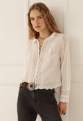 A woman wearing a white long-sleeve blouse with intricate lace and embroidered detailing, tucked into dark gray jeans with a brown belt, standing against a white paneled wall.