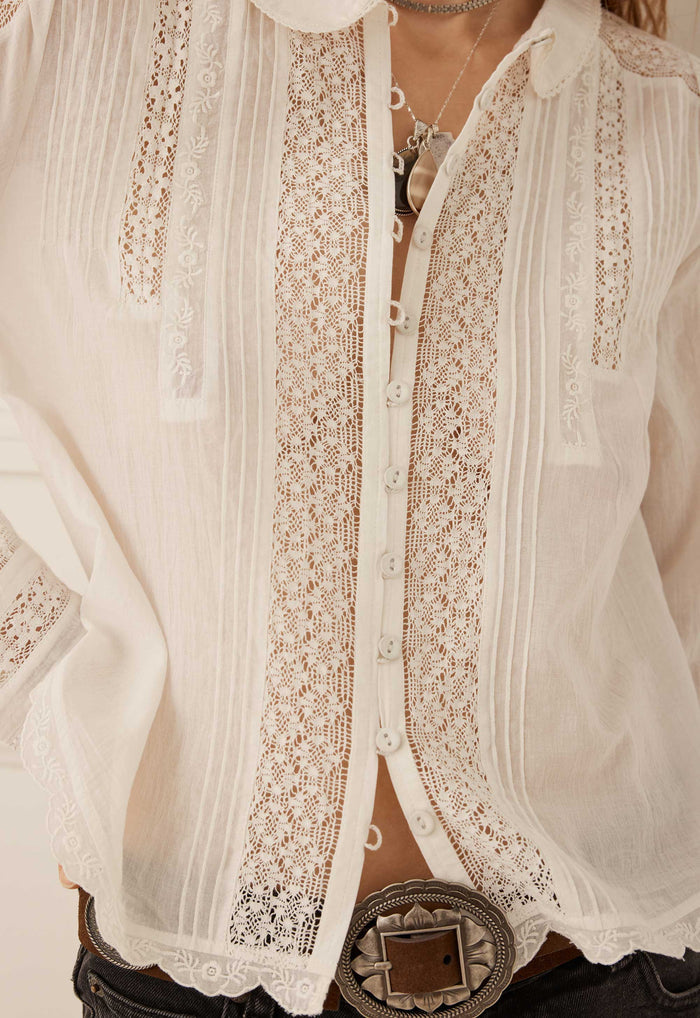 A close-up of a white long-sleeve blouse with intricate lace panels and vertical pleated detailing, styled with a large silver concho belt and necklace.