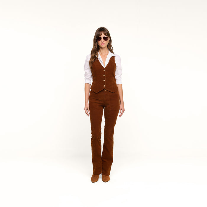 A woman stands against a white background wearing sunglasses, a white shirt, a brown vest, and the Saffia Corduroy Pant with boots.