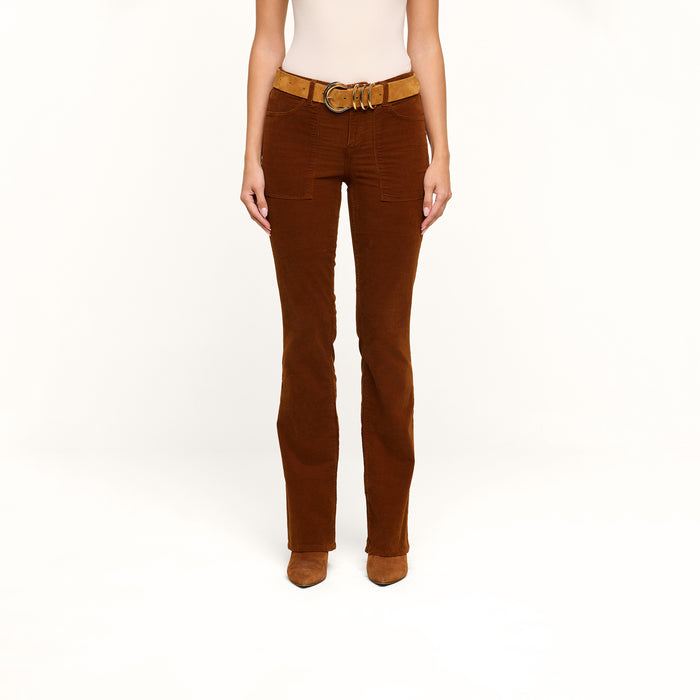 Model showcases the lower half of their body wearing the Saffia Corduroy Pant in brown, styled with a tan belt and brown shoes, posed against a plain white background.