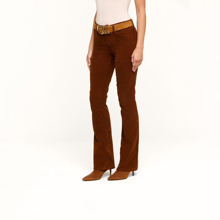 A person is wearing the Saffia Corduroy Pant, a high rise brown flared pant made from stretch corduroy, styled with a tan belt and brown pointed shoes, standing against a plain white background. The upper body is partially visible.