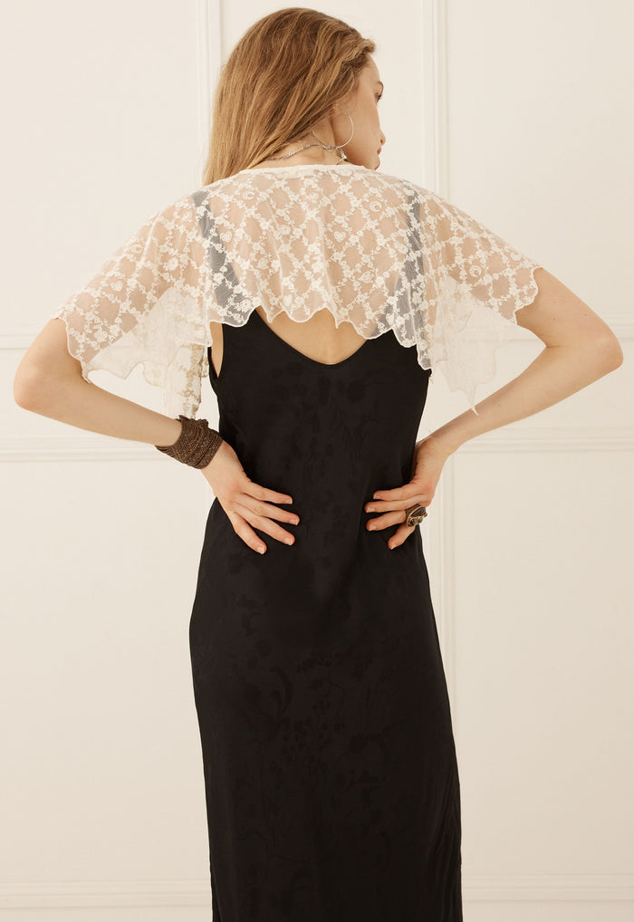 A woman viewed from the back wearing a black slip dress with a sheer white lace flutter cape overlay, set against a light cream paneled wall.