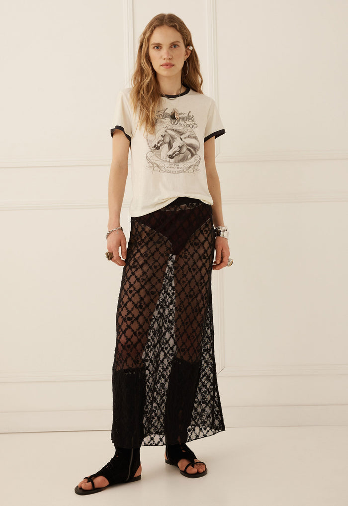 A model stands against a white paneled wall wearing a cream graphic t-shirt with black trim, sheer black patterned maxi pants, and black sandals.