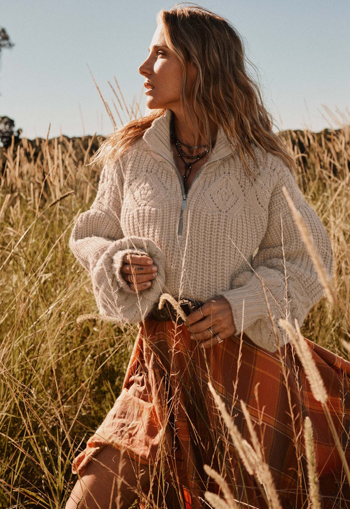 A woman standing in a field of dry golden grass, wearing a cream cable-knit half-zip sweater and a rust-orange plaid skirt.