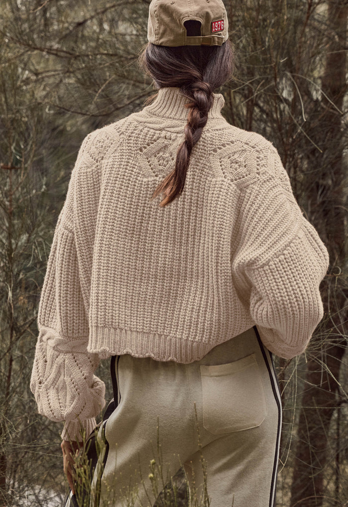 A person shown from behind wearing a cream-colored chunky knit cropped sweater, beige pants, and a beige cap, standing outdoors in a wooded setting.