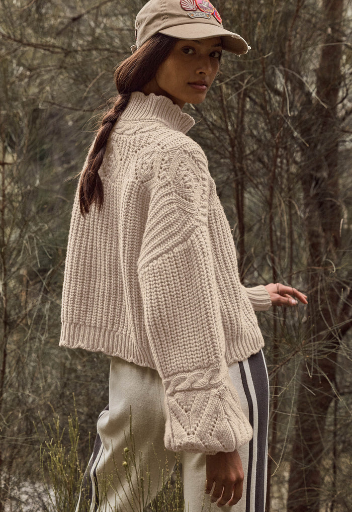 A woman wearing a chunky cream cable-knit mock-neck sweater, a beige baseball cap, and cream pants with black stripes, standing in a wooded outdoor setting.