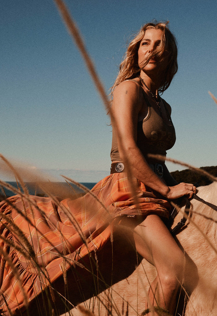 A woman outdoors in a desert landscape wearing a brown halter top and an orange plaid skirt, seated on a horse with dry grass in the foreground.