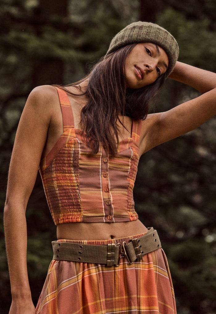 A woman wearing a rust-orange and brown plaid sleeveless crop top with button details, a matching plaid skirt, and a textured olive green beanie, photographed outdoors against a blurred forest background.