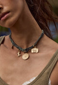 A close-up of a woman wearing a dark olive green braided leather choker necklace with gold-toned animal and circular pendant charms.