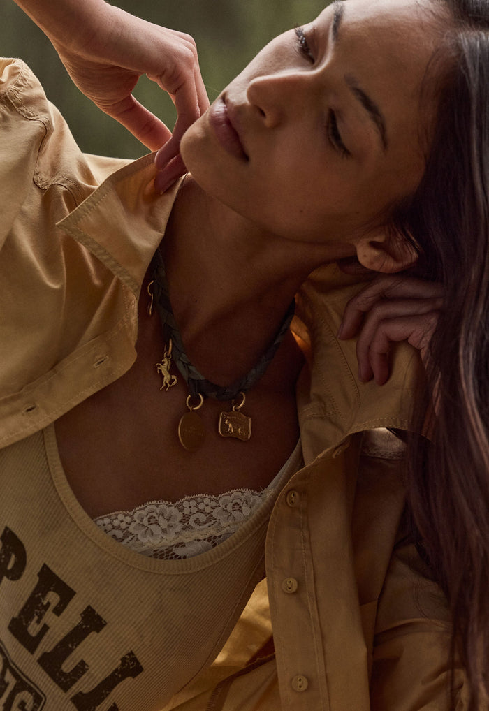 A close-up lifestyle shot of a woman wearing a layered necklace set. The set includes a dark green braided cord necklace with a small gold charms.
