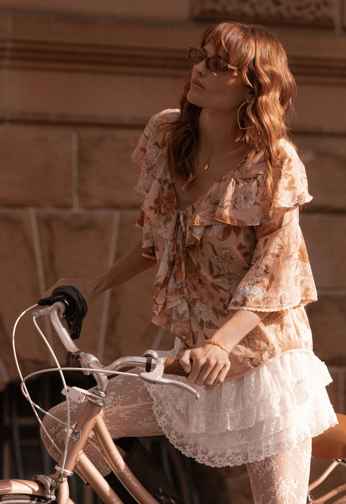 Model beside a bicycle, front ruffle and tie detail of Rose Quartz floral organic cotton blouse
