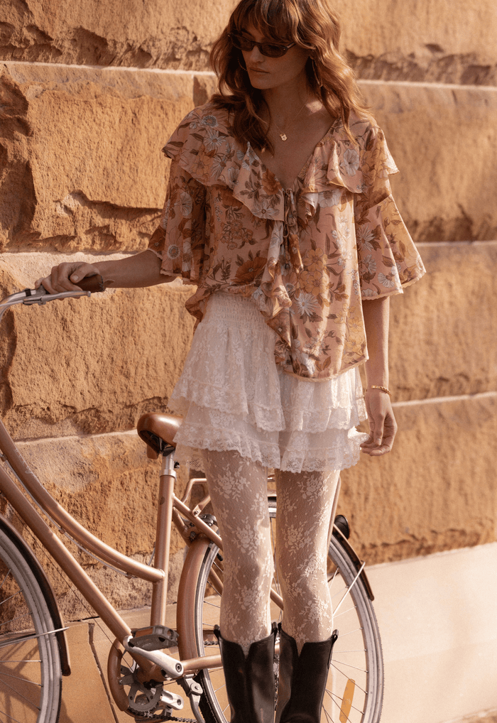 Model wearing Stealing Beauty blouse in Rose Quartz with tiered ruffles and front ties, styled with lace skirt and boots