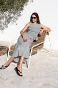 A woman wearing a sleeveless gray midi dress and black sunglasses sits on a woven outdoor lounge chair against a white wall.
