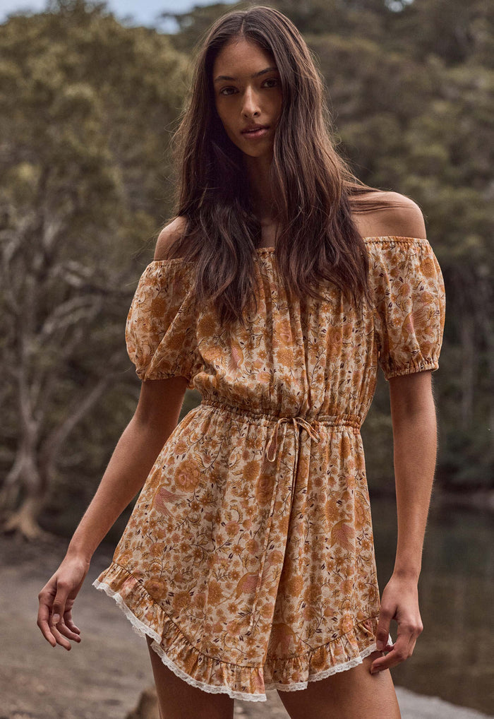 A woman wearing a short, off-the-shoulder dress with puff sleeves and a gathered waist, featuring an orange and beige floral pattern. She is standing outdoors in front of a blurred natural background of trees.