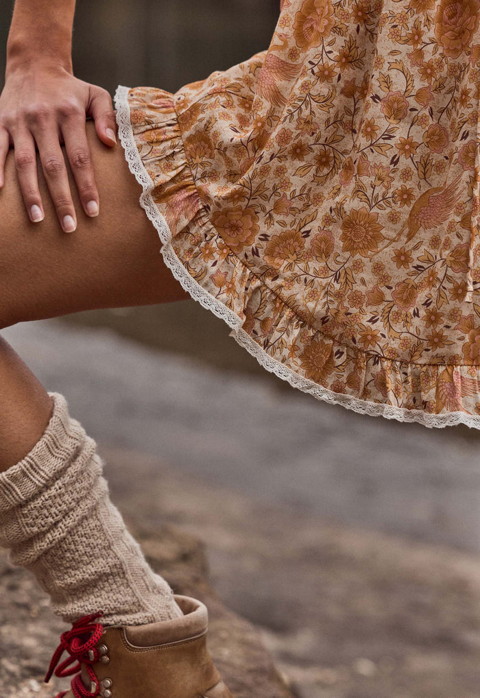 Close-up of a person wearing a tan and brown floral skirt with a white lace hem, paired with beige knit leg warmers and brown boots with red laces.