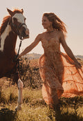 A woman in a flowing rust-orange boho dress walks beside a brown and white horse in an outdoor grassy field at golden hour.