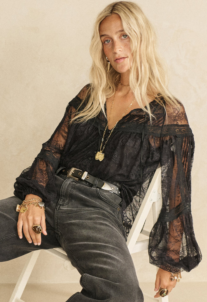 Model seated front-on showing elastic cuffs and pintuck detail on black lace blouse, styled with grey jeans and western belt
