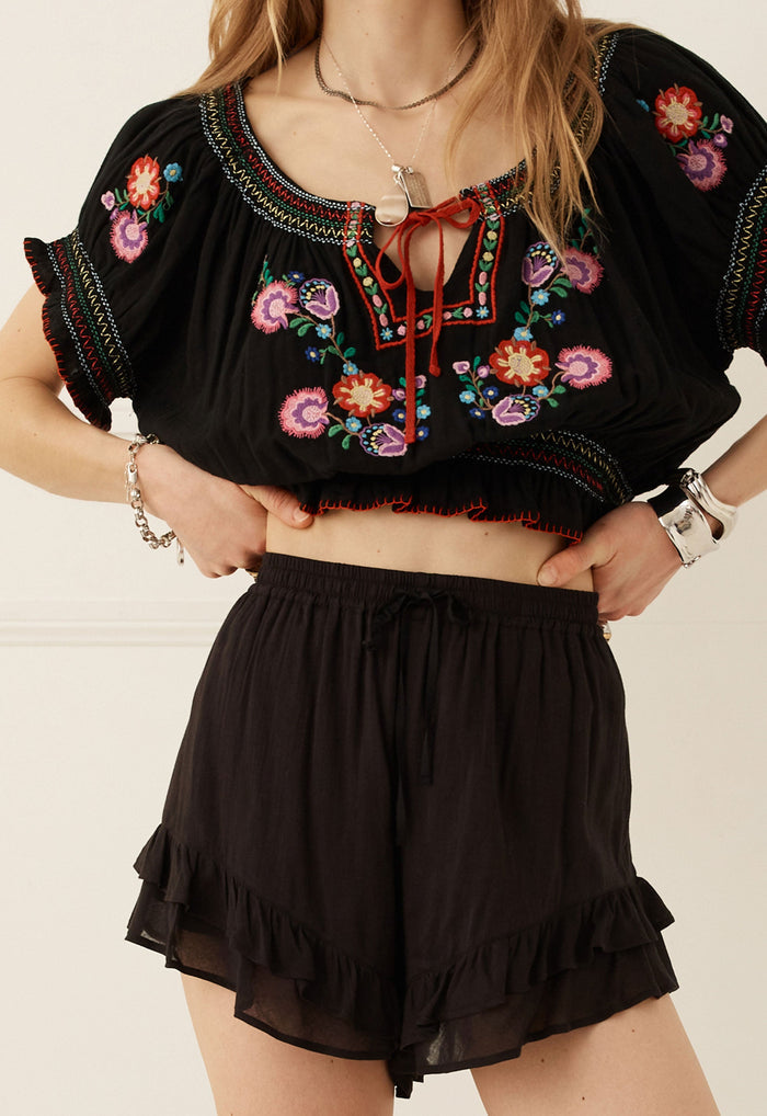 A woman wearing a black embroidered crop top with colorful floral patterns and matching black ruffled shorts, standing against a light beige wall.