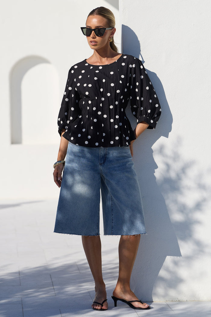 A woman stands outdoors against a white wall wearing a black long-sleeve blouse with white polka dots, light-wash blue denim culottes, and black sandals.