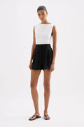 A model wearing a sleeveless white top and black shorts, standing against a plain white studio background.