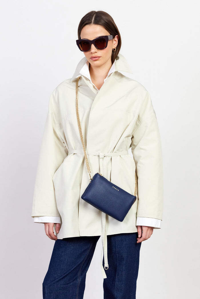 Woman wearing a cream coat and dark sunglasses, holding a navy blue clutch against a white background