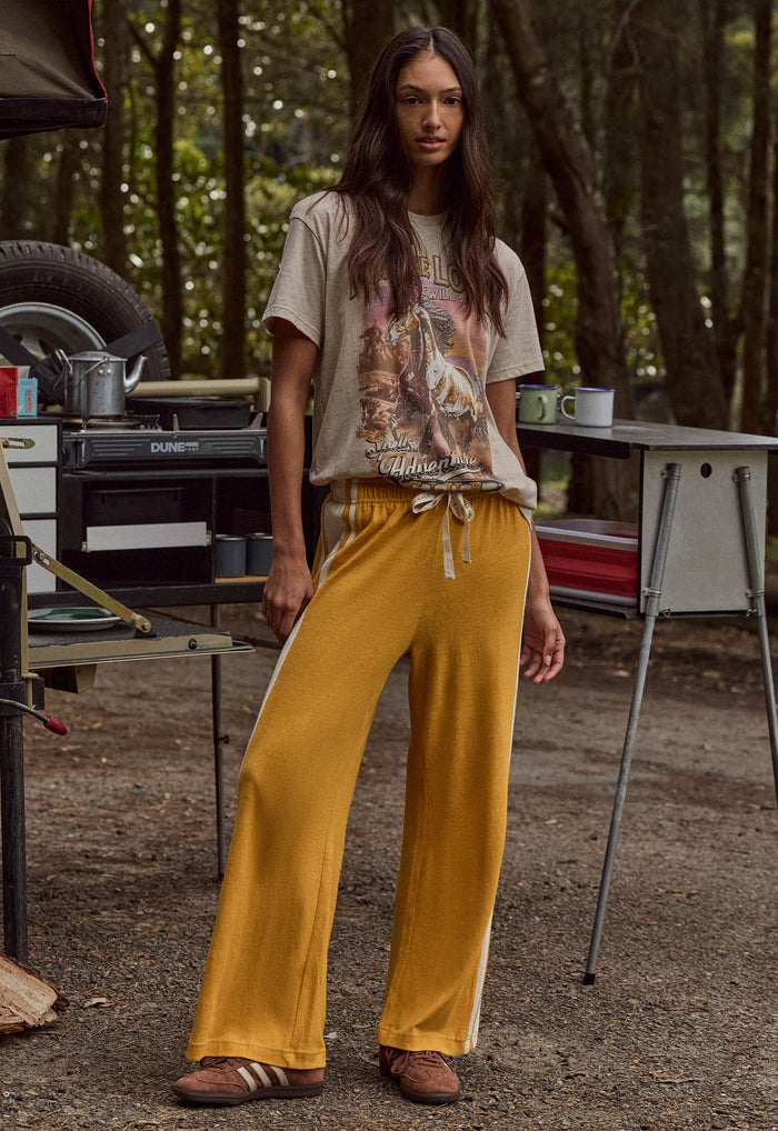 A woman stands outdoors in a wooded campsite setting wearing loose mustard yellow drawstring pants with white side stripes, a beige graphic T-shirt, and brown shoes.