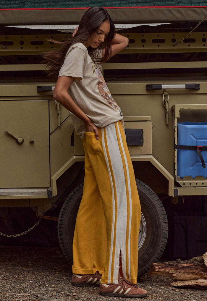 A woman stands outdoors in front of a military-style vehicle, wearing a beige graphic T-shirt, mustard yellow track pants with white side stripes, and brown sandals.
