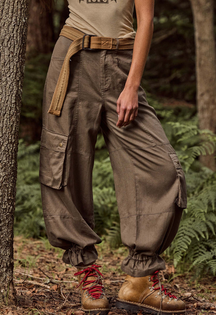 A person standing outdoors in a forest wearing loose brown cargo pants with gathered cuffs, a tan belt, and brown lace-up boots.