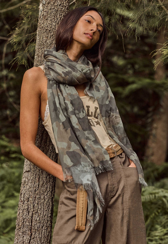 A woman in a forest setting wearing a camouflage print scarf draped over her shoulders, paired with a beige tank top and brown cargo pants.