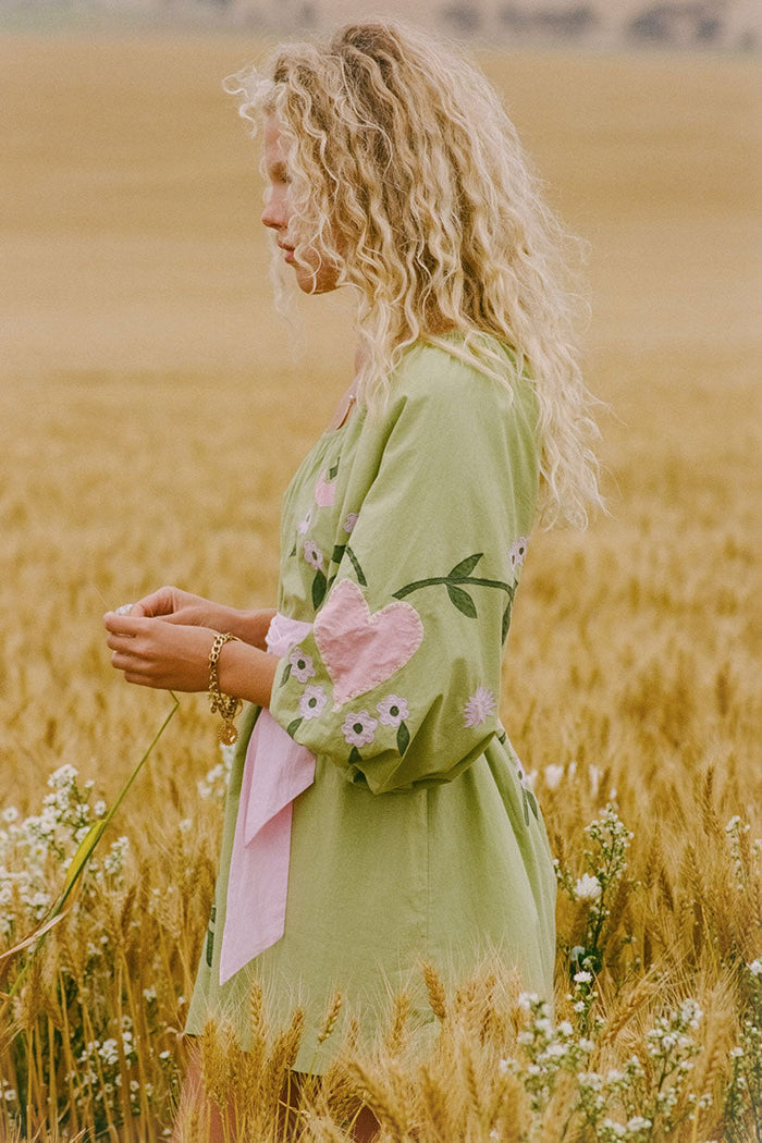 Side profile view of Wonderland Dress in Lime Essence with pink heart appliqué, floral details, and pink bow tie waist