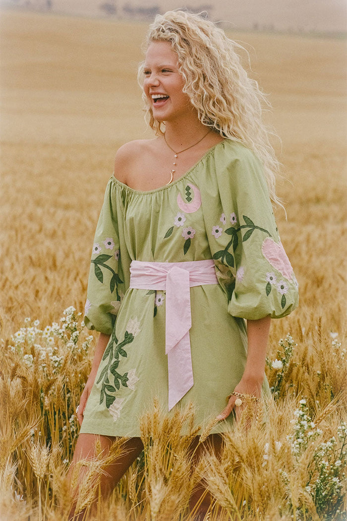 Front view, sage green organic cotton mini dress with pink floral appliqué, puff sleeves, elastic neck, and pink sash belt