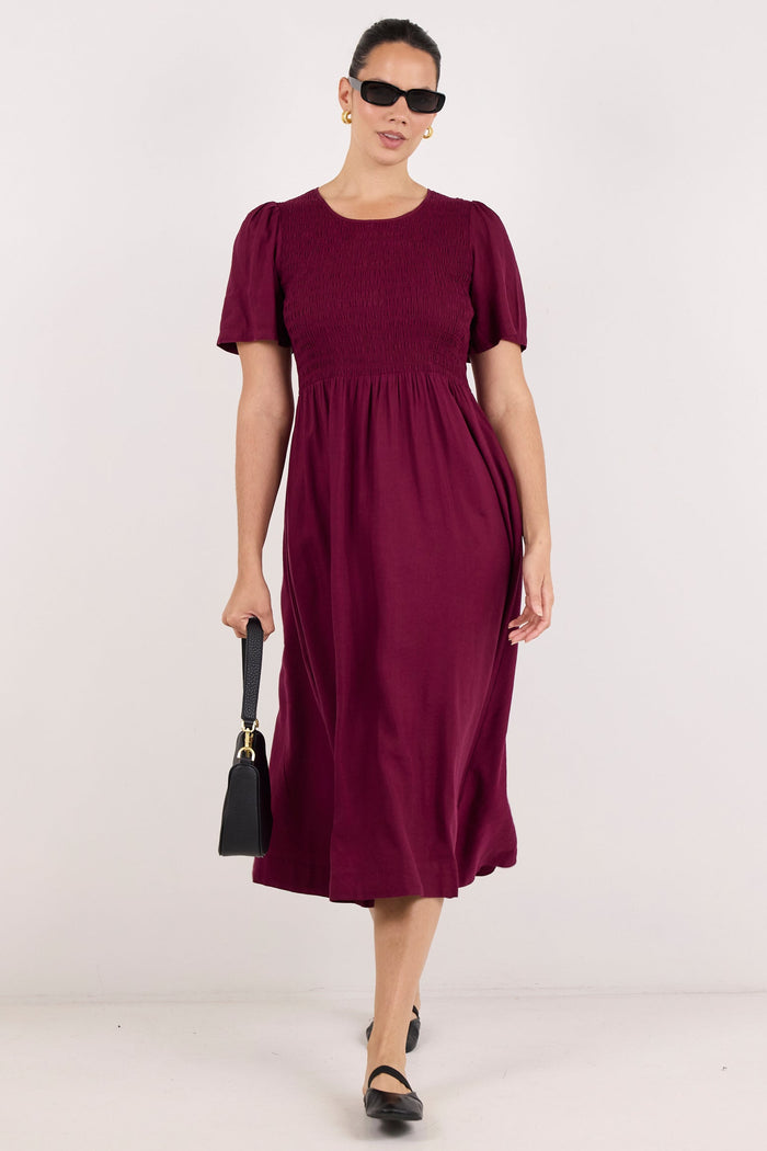 A woman modeling a short-sleeve midi dress in deep burgundy, paired with black sunglasses, black shoes, and a small black handbag, set against a plain white studio background.