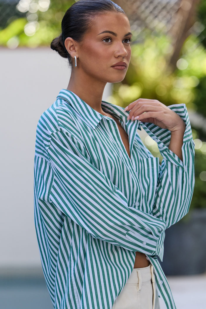 A woman wearing an oversized green and white striped button-down shirt with cream pants, photographed outdoors with a blurred background.