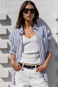 A woman wearing a navy and white vertically striped button-down shirt over a white tank top, paired with white pants and black sunglasses, posing against a white brick wall.
